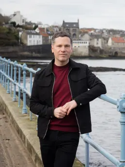 Allan Radcliffe leaning against a pale blue harbour railing in front of a Scottish coastal village
